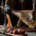 Whole Iberian ham on stand with bread, tomatoes and olive oil on rustic table inside a traditional curing cellar.