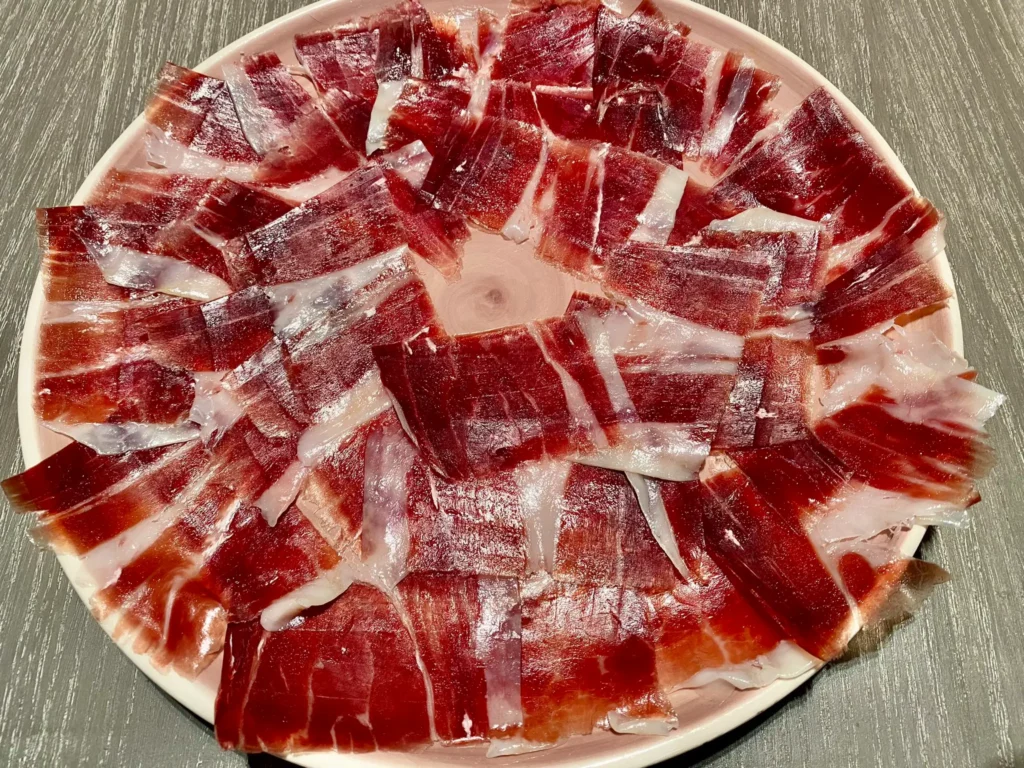 Thin slices of Iberian ham arranged in a circular pattern on a plate, showcasing its marbling and deep red color
