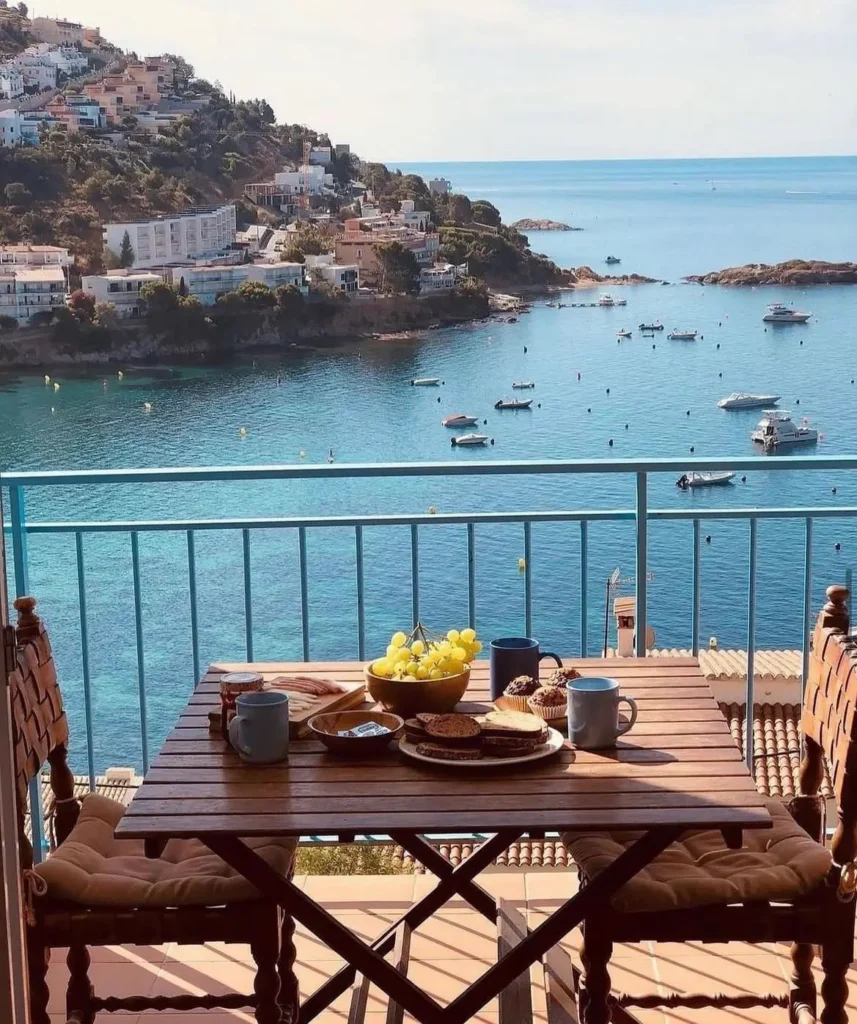 Breakfast table on a Mediterranean balcony overlooking the sea with boats and hillside houses in the background
